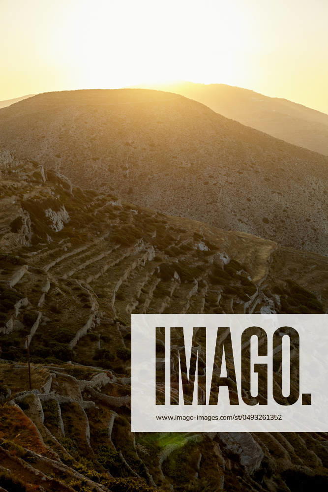 The sun sets behind a hill, casting a warm glow over terraced slopes with a network of paths or steps carved into the landscape. Folegandros Greece bola01002