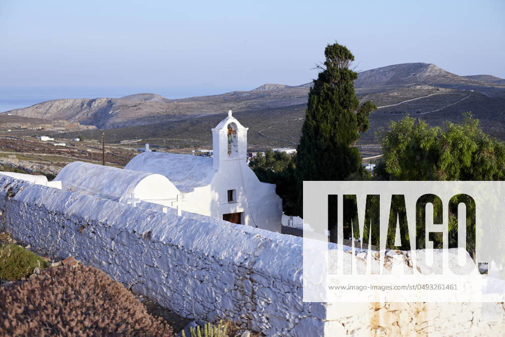 A serene landscape featuring a traditional white-washed church with a bell tower, surrounded by a stone wall, overlooking a hilly terrain with the sea in the distance at dawn. Folegandros Greece bola01005