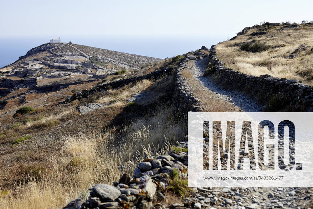 A stone hiking trail leads through a dry, grassy landscape toward a distant overlook with a clear blue sky above. Folegandros Greece bola01011