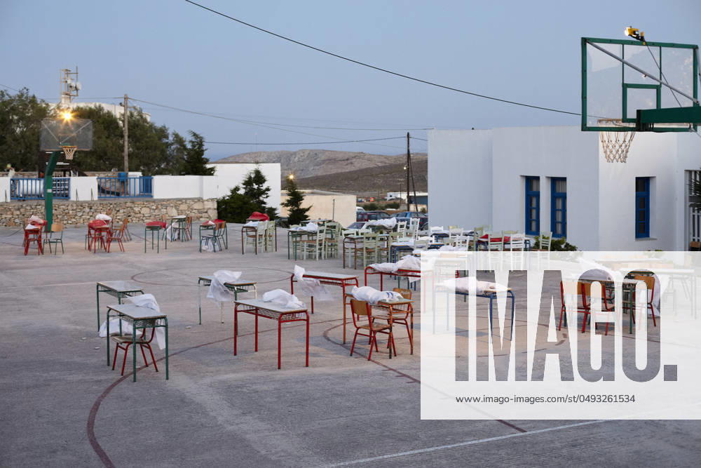 An outdoor setting at dusk featuring a basketball court with tables and chairs neatly arranged across it, under the glow of gentle lights, adjacent to a white building with blue trimmings. Folegandros Greece bola01016