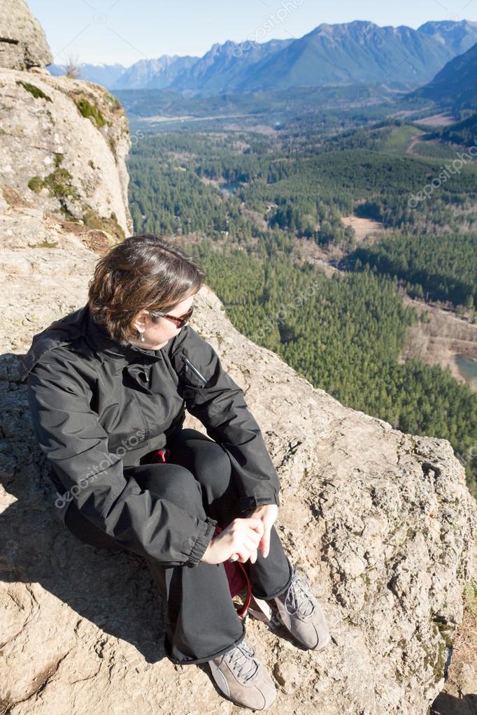 Young woman wearing sunglasses sitting on a rocky outcrop enjoying the view of the forested valley, lake and mountains from Rattlesnake Ledge Trail, Snoqualmie, Washington after hiking up the summit model released