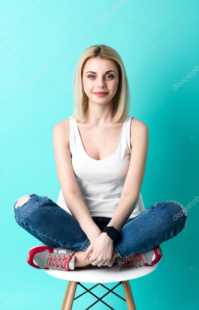 Portrait of attractive young woman sitting on chair in lotus position with comfort. She is looking at camera with joy and smiling. Isolated on blue background model released