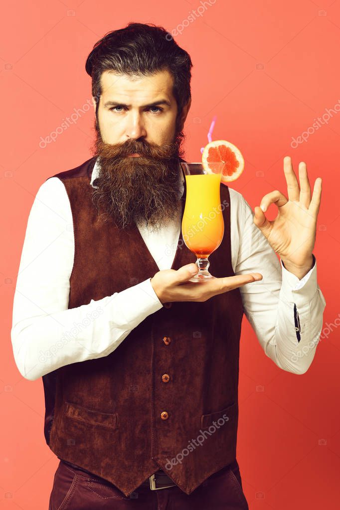 Handsome bearded man with long beard and mustache has stylish hair on serious face holding glass of alcoholic cocktail in vintage suede leather waistcoat on red studio background. model released