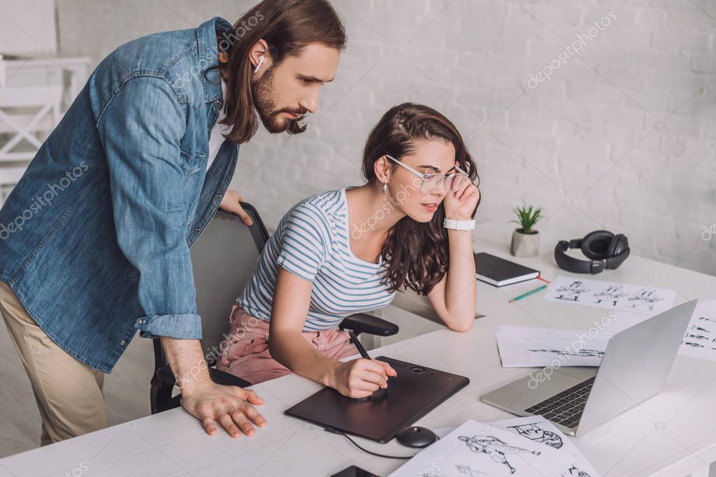Illustrator looking at laptop near coworker in glasses and sketches model released