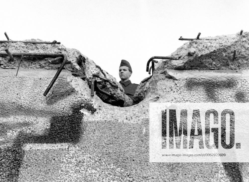 Soldat der Grenztruppen schaut über teilzerstörte Berliner Mauer - iblzni01084378