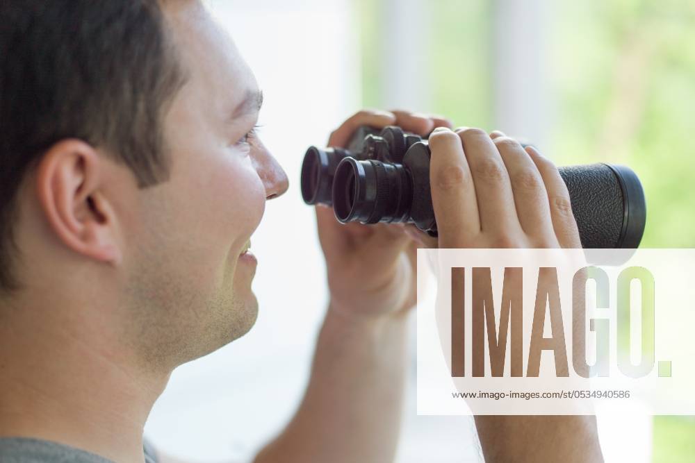 Young man standing looking through a glass window with binoculars as he watches something in the distance ,model released, Symbolfoto; Jung Mann stehend Schauen durch einer Glas Fenster mit Fernglas als er Uhren etwas in der Entfernung ,model released, Symbolfoto ING_32193_135869