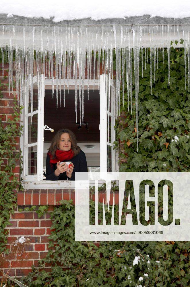 Junge Frau wärmt sich an einem Becher und schaut im Winter aus dem Fenster ihres Hauses, an dem lange Eiszapfen hängen iblbcc01445766