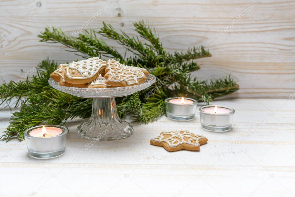 Gingerbread cookies for Christmas on a crystal glass bowl, burning candles and fir branches on a white painted wooden table, copy space, selected focus