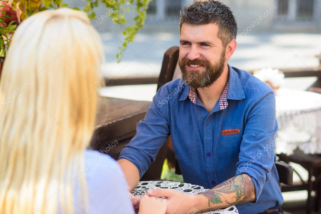 Man with happy face fall in love with blonde, look, gaze at her. Couple in love sit at terrace, cafe outdoor, urban background, defocused. Fall in love concept. Couple speaking, hang out, have fun. model released