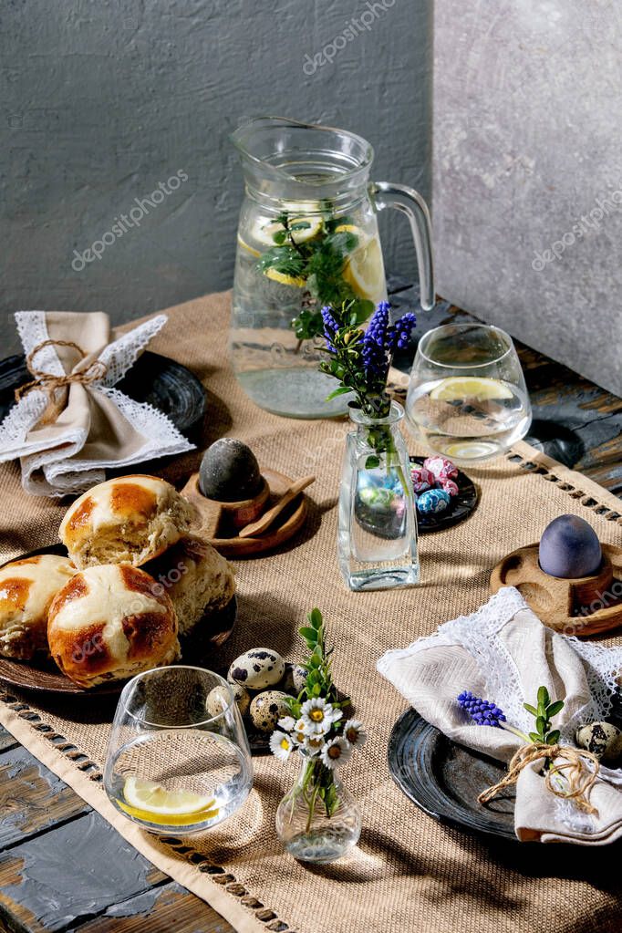 Easter table setting with colored and chocolate eggs, hot cross buns, bouquet flowers, empty ceramic plate with napkin, glass of lemonade drink on wooden table with textile tablecloth.