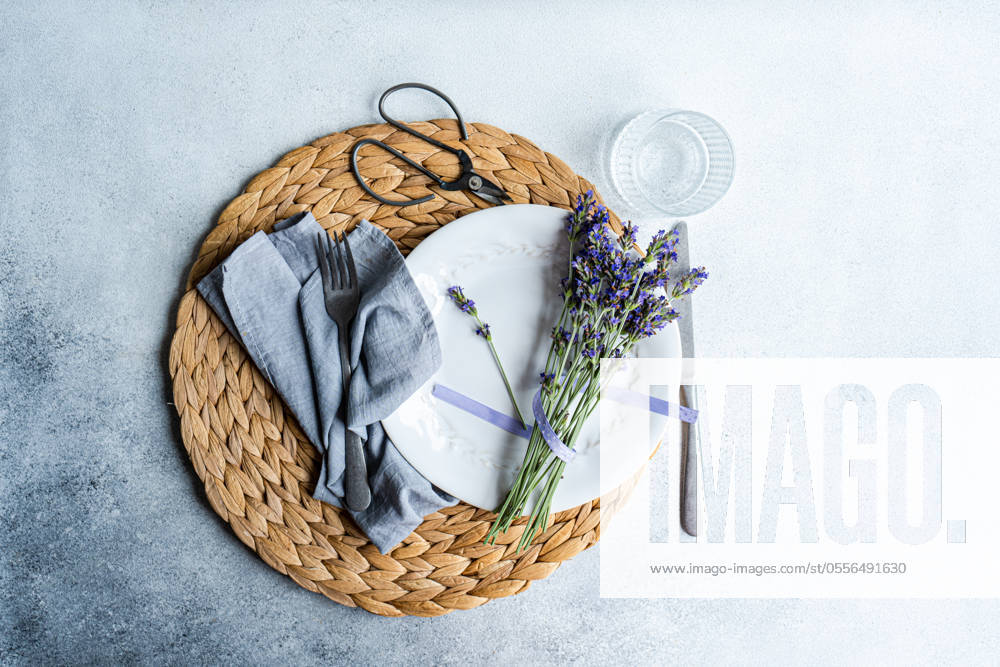 An inviting summer table setting featuring a woven placemat, white dinnerware, linen napkin, cutlery, a glass, and fresh lavender flowers Model Released AnnaBogush_ID21818_639754_004