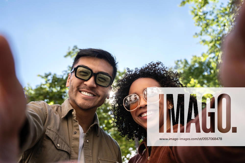 IMAGO Creative: Sommer, Sommertage im Stadtpark Multiethnic couple taking a selfie in a sunny Madrid park. The Chinese man and Hispanic woman are smiling, wearing sunglasses, and enjoying the outdoors. The background features greenery and clear blue sky. Model Released ManuelRuiz_ID21992_644068_043