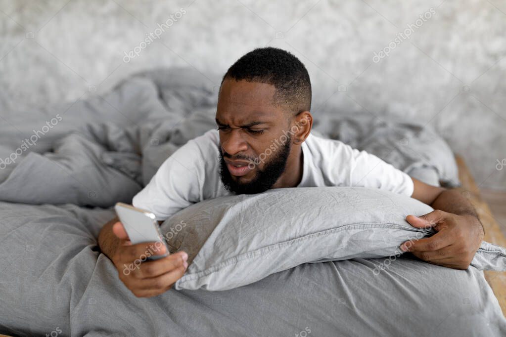 Oversleep Concept. Portrait of confused black man waking up with anxiety, upset angry young guy turned off alarm clock on smartphone lying on bed in bedroom, reading bad news, late for work model released