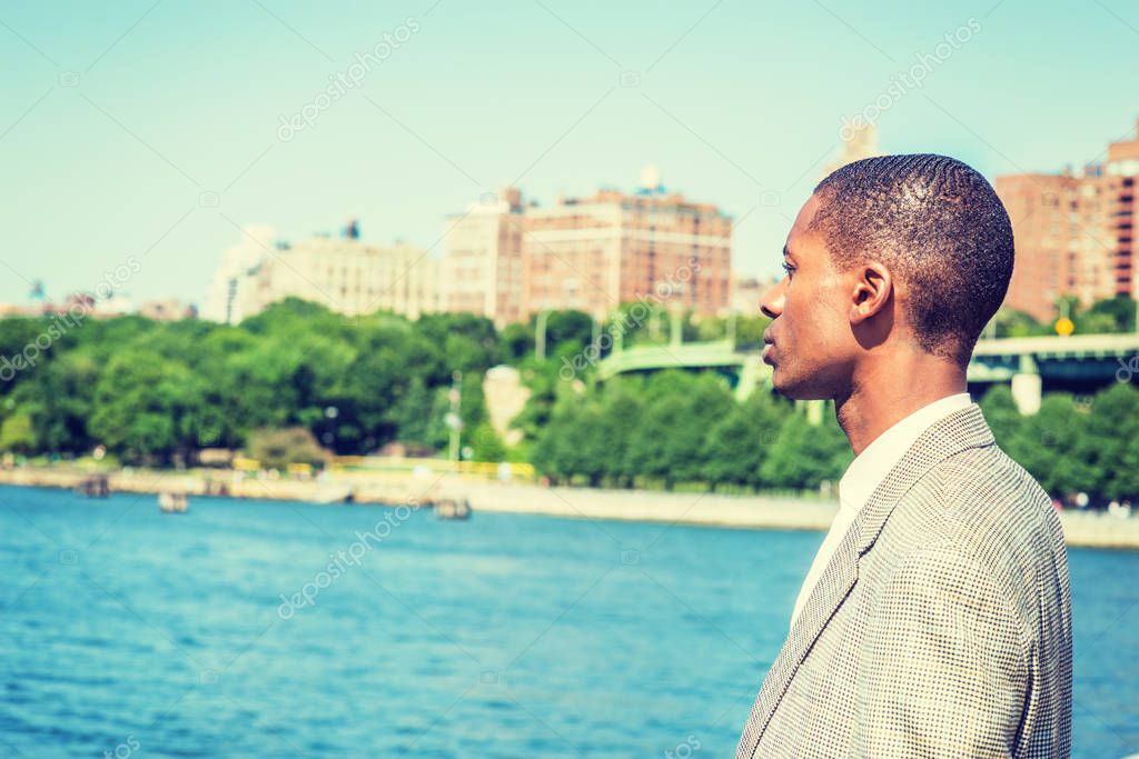 Young African American Man with short hair, traveling in New York City, standing by Hudson River in Manhattan under sun, looking, waiting, thinking. Green trees, buildings on background. Side View model released