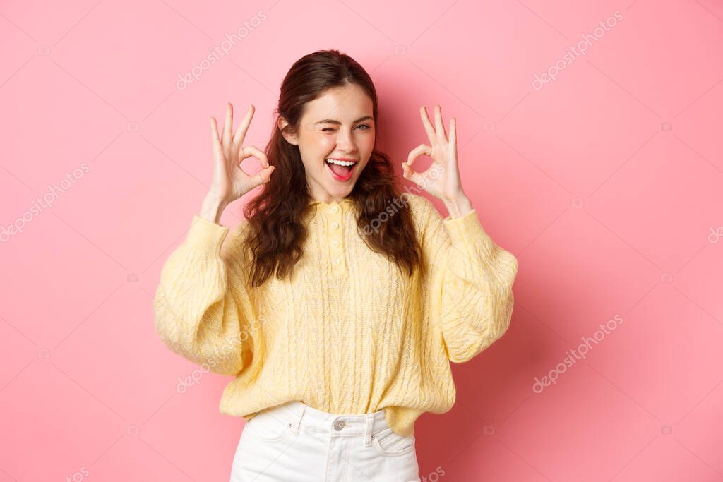 Portrait of cheerful pretty girl winking and saying yes, showing okay sign, alright gesture, praise good job, well done, standing pleased against pink background. model released