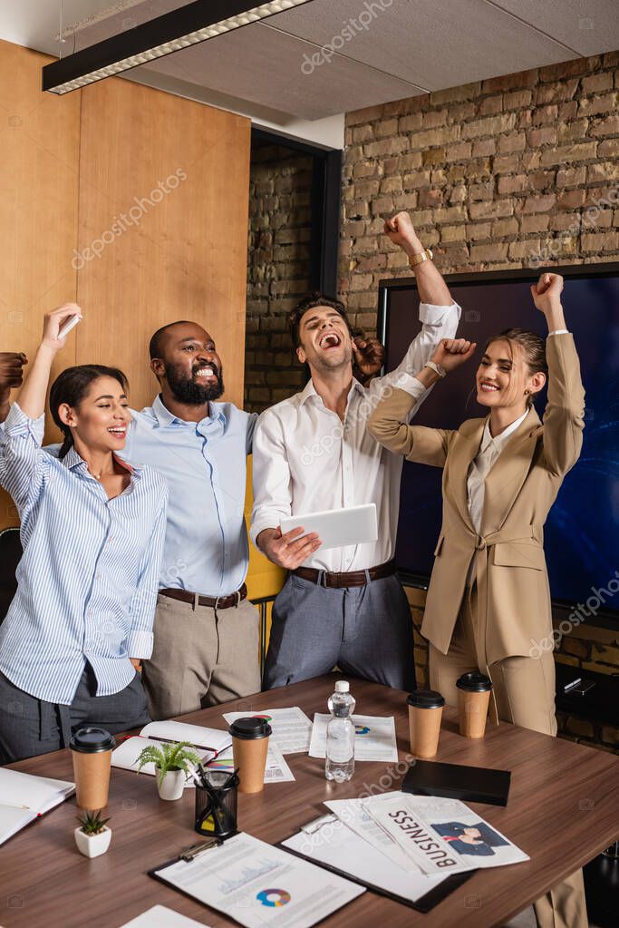 Excited businessman holding digital tablet near cheerful interracial colleagues showing win gesture model released