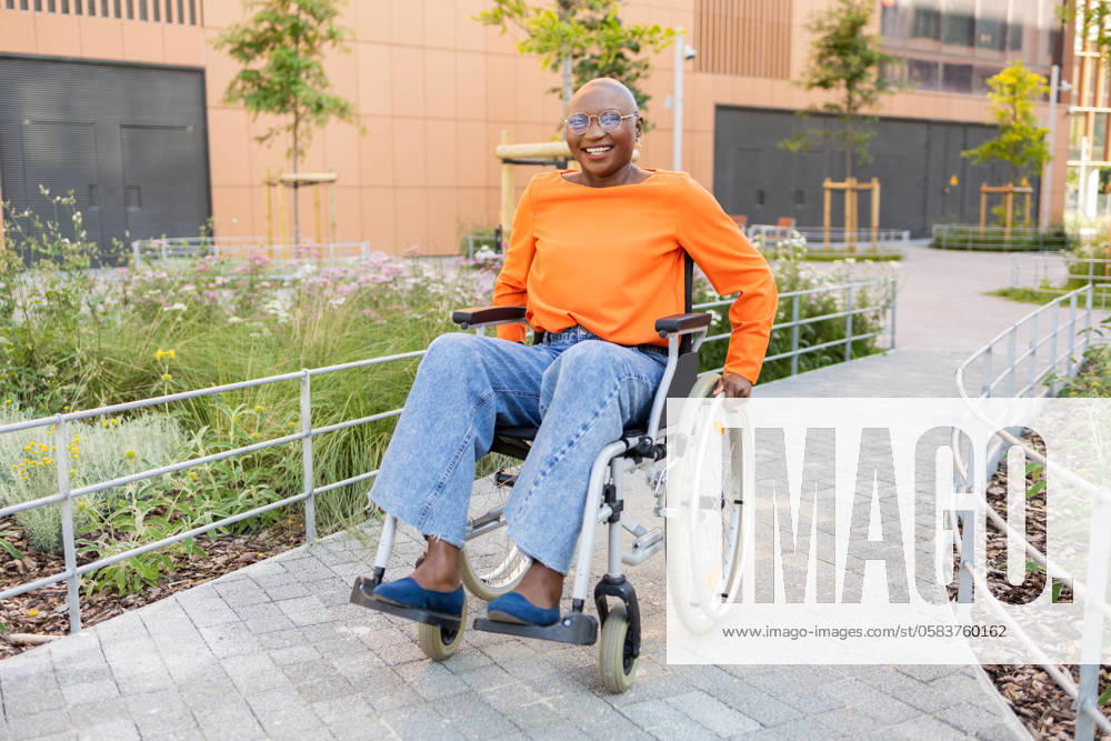 IMAGO Creative: Sommer, Sommertage im Stadtpark Black woman with a bright smile, dressed in orange top and jeans, navigates a wheelchair through a serene park, exemplifying accessibility and joy Model Released DavidMunoz_ID22272_651064_052