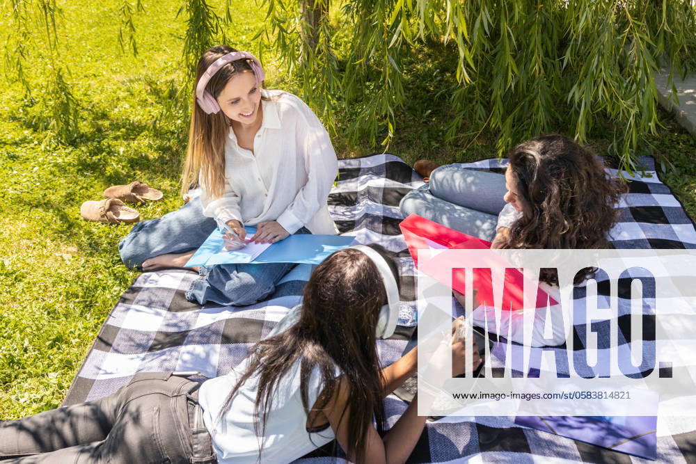 IMAGO Creative: Sommer, Sommertage im Stadtpark Three multiethnic women engage in a productive outdoor meeting at a park, sitting on a plaid blanket under the shade of weeping willows, reviewing documents and using digital devices Model Released IgnacioFerrandiz_ID22117_647165_038