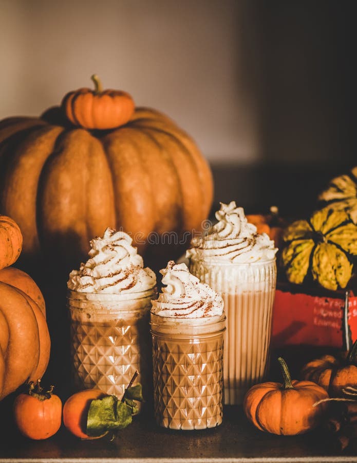 Pumpkin latte coffee with whipped cream in glasses on table, Pumpkin spice latte coffee drink topped with whipped cream and cinnamon in tall glasses among fresh pumpkins and persimmons on table, grey wall at background. Seasonal Autumn hot warming sweet drink