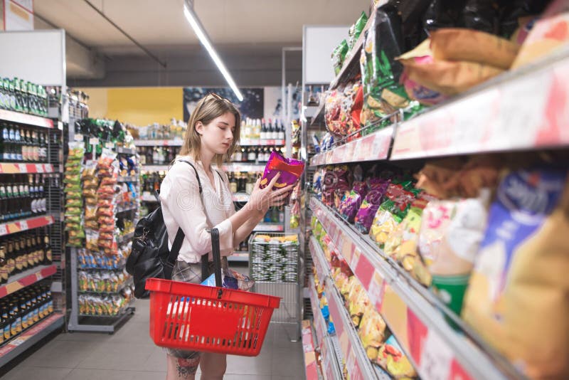 Girl with a red basket in her hands chooses chips from the shelves of the supermarket, Girl with a red basket in her hands chooses chips from the shelves of the supermarket. Attractive young woman buys chips in the snack department at the supermarket. Shopping in a supermarket concept model released