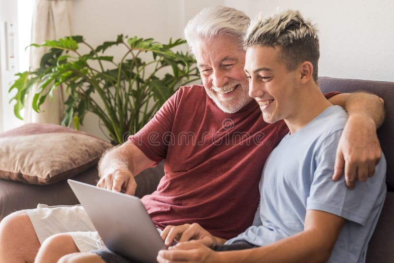 Grandfather and grandson sitting together on the sofa at home in friendship looking at the same laptop and smiling - sharing the, Grandfather and grandson sitting together on the sofa at home in friendship looking at the same laptop and smiling - sharing the same interest or passion. Concept of family love