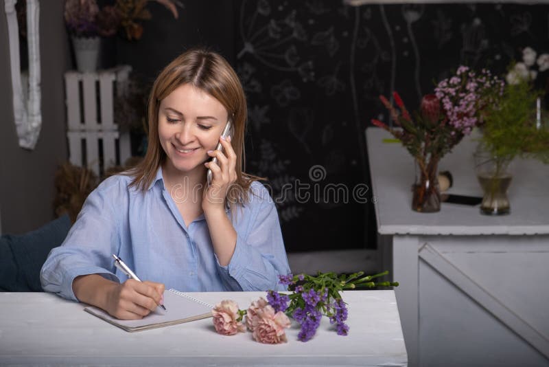 Smiling young female florist standing in her flower shop using her telephone and notebook to take orders for her store., Smiling young female florist standing in her flower shop using her telephone and notebook to take orders for her store model released