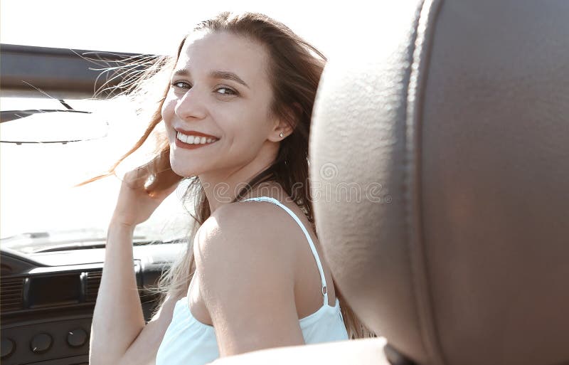 Happy woman sitting on passenger seat in car, Back view of joyful young female in summer clothes sitting on passenger seat of cabriolet and looking at camera over shoulder during road trip model released