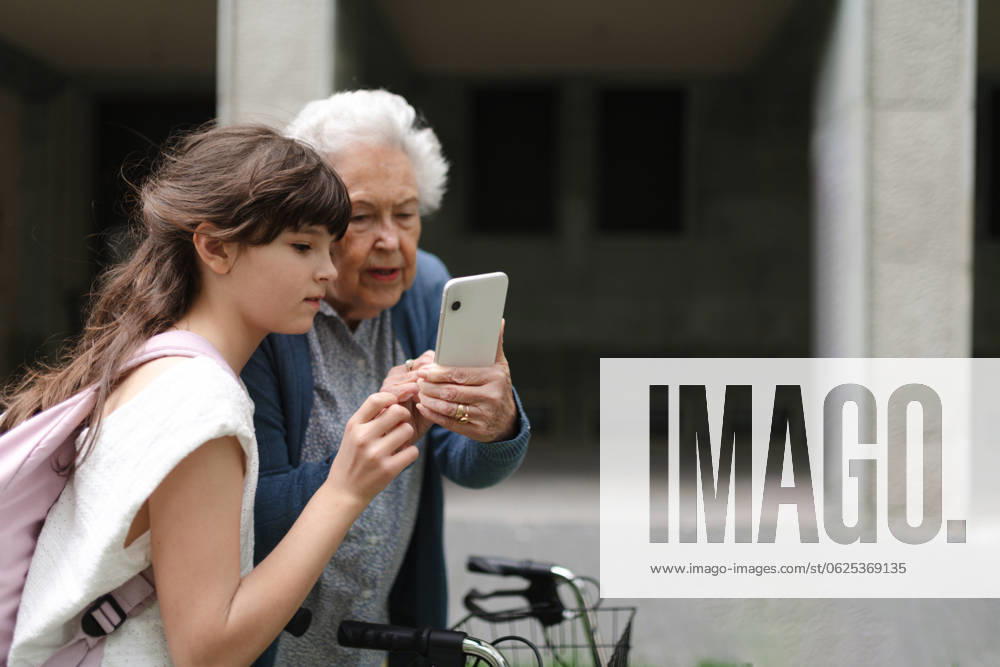 Grandma picking up young girl from school at afternoon. Granddaughter showing something on smartphone to senior grandmother, teaching her how to work with technology. Grandma picking up young girl from school at afternoon. Granddaughter showing something on smartphone to senior grandmother, teaching her how to work with modern technology. model released