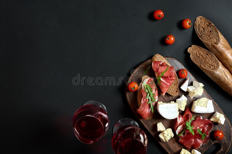 Prosciutto, salami, baguette slices, tomatoes and nutson rustic wooden board, two glasses of red wine over black, Cheese and meat appetizer selection. Prosciutto, salami, baguette slices, tomatoes and nutson rustic wooden board, two glasses of red wine over black background. Still life