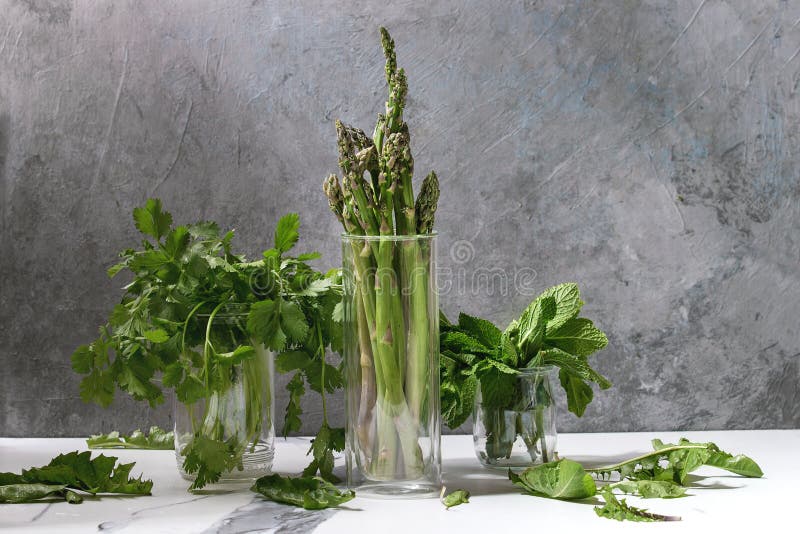 Asparagus ang greens, Raw uncooked organic green asparagus, bunch of coriander herbs and fresh mint in glass jars on white kitchen table with grey texture wall as background. Copy space.