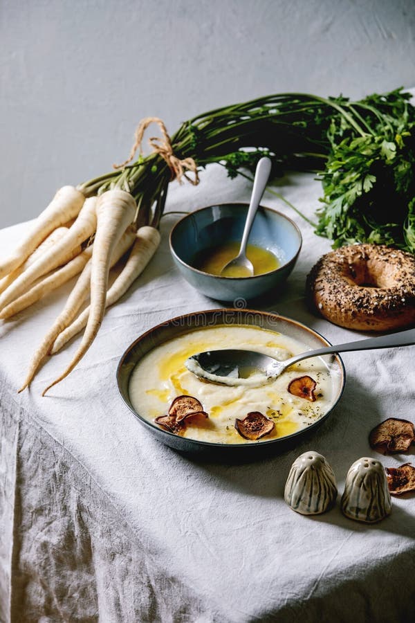 Parsnip soup with butter sauce, Parsnip cream soup in ceramic bowl with butter sauce, sun dried pears, bundle of fresh parsnip, bagel bread and herbs on kitchen table with white tablecloth