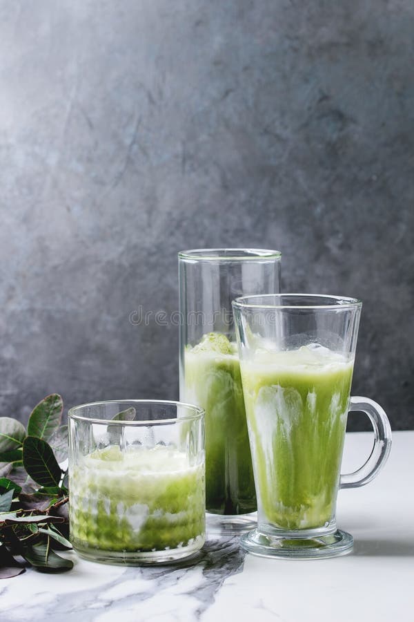 Matcha iced latte, Matcha green tea iced latte or cocktail in three different glasses with ice cubes, matcha powder and jug of milk on white marble table, decorated by green branches. Grey wall at background
