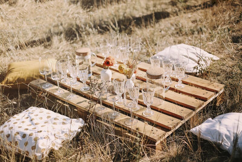 Wooden table with glassware arranged for picnic, Set of elegant glassware placed on plank wooden table with floral arrangement on grassy meadow during festive autumn picnic