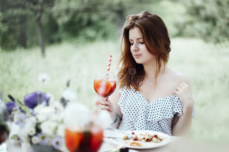 Cheerful woman with cocktail during banquet, Happy young female with alcohol cocktail smiling and proposing toast while sitting at table during banquet in garden model released