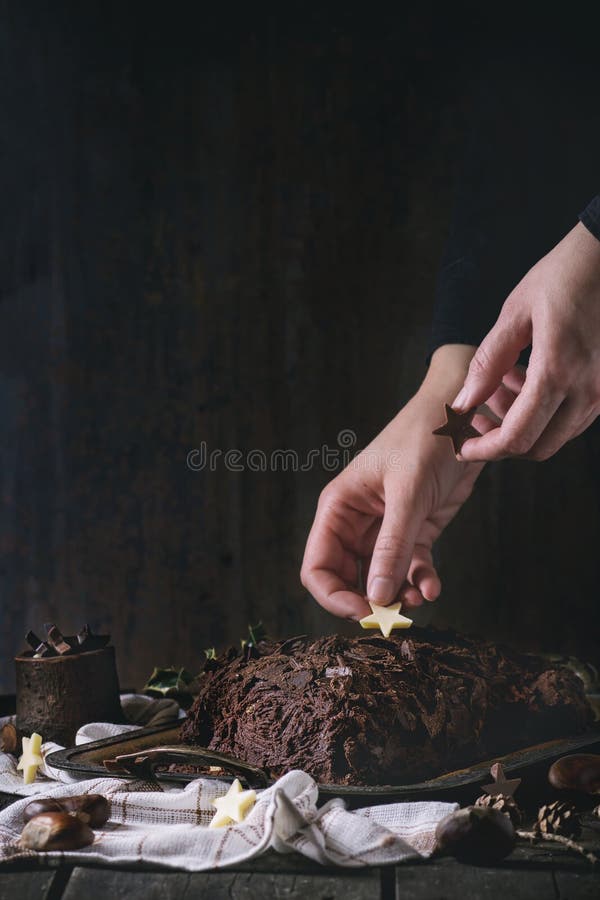 Decorating of christmas chocolate yule log, Decorating process of homemade Christmas chocolate yule log by womans hands with chocolate stars over old wooden table with holly branch and chestnuts. Dark rustic style. With retro filter effect
