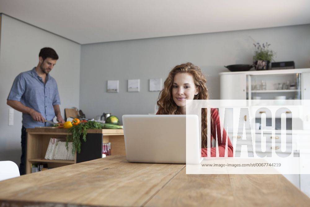 Woman at home using laptop with man in background cutting vegetables model released Symbolfoto property released RBF003543