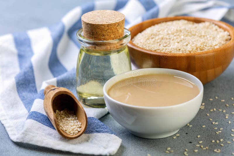 Bottled sesame oil, tahini and white sesame seeds, Cold pressed sesame oil from white sesame, tahini and sesame seeds in wooden bowl on grey textured background, selective focus