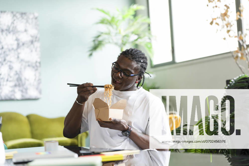 A black man sits in a comfortable indoor setting, using chopsticks to eat noodles from a takeout box Model Released VictorCuenca_ID22085_646425_028