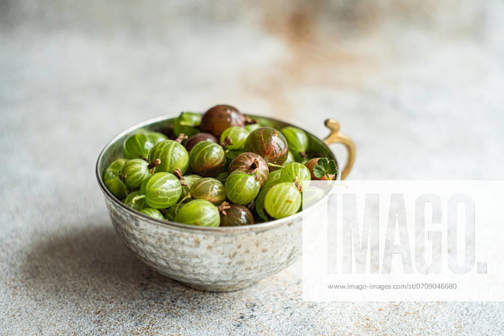 From above, a variety of fresh organic gooseberries in hues of green and burgundy rest in an ornate metal bowl on a textured grey surface, with subtle lighting enhancing their natural sheen. Model Released AnnaBogush_ID22657_660678_004