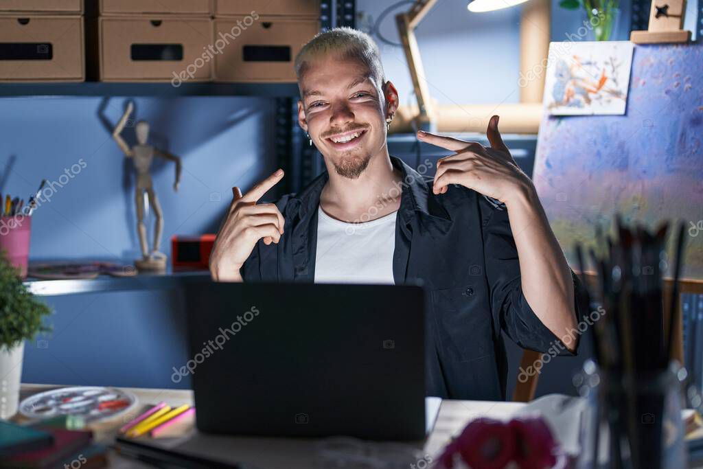 Young caucasian man using laptop at night at art studio smiling cheerful showing and pointing with fingers teeth and mouth. dental health concept.