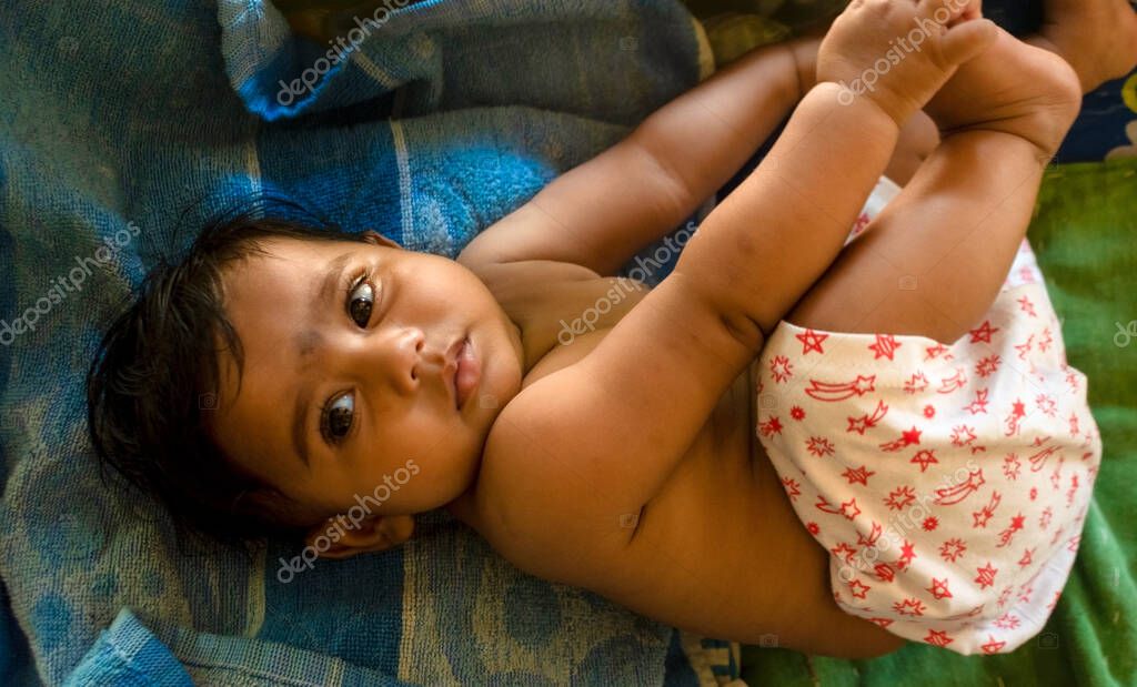 Cute indian baby lying on a blue towel holding her feet looking away from camera. Selective focus on subject's left eye with background blur.