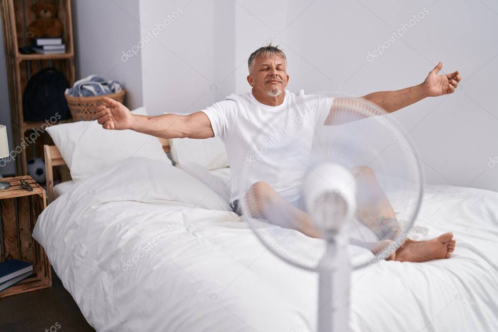 Middle age grey-haired man using ventilator sitting on bed at bedroom