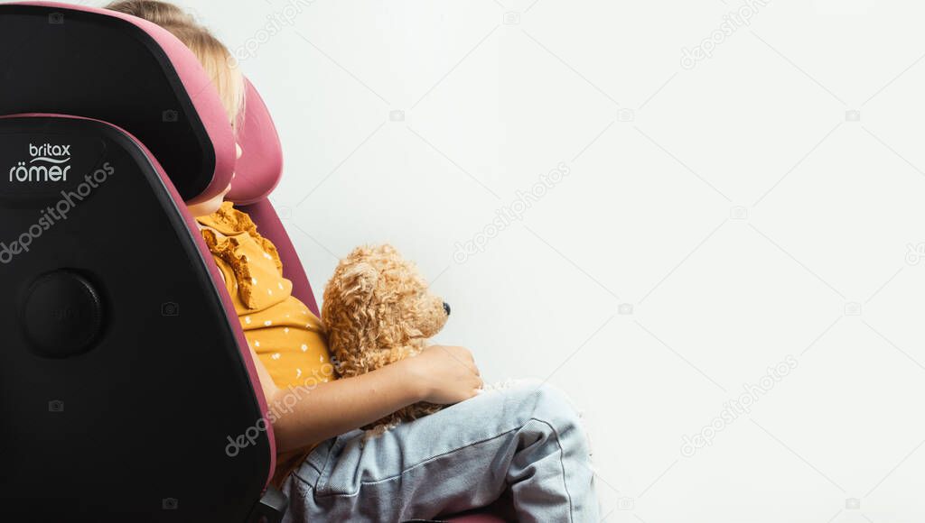 Cute little caucasian girl fastened with a belt in a Britax Romer safety car seat with sict isolated on white background. Kid safety while traveling by car.