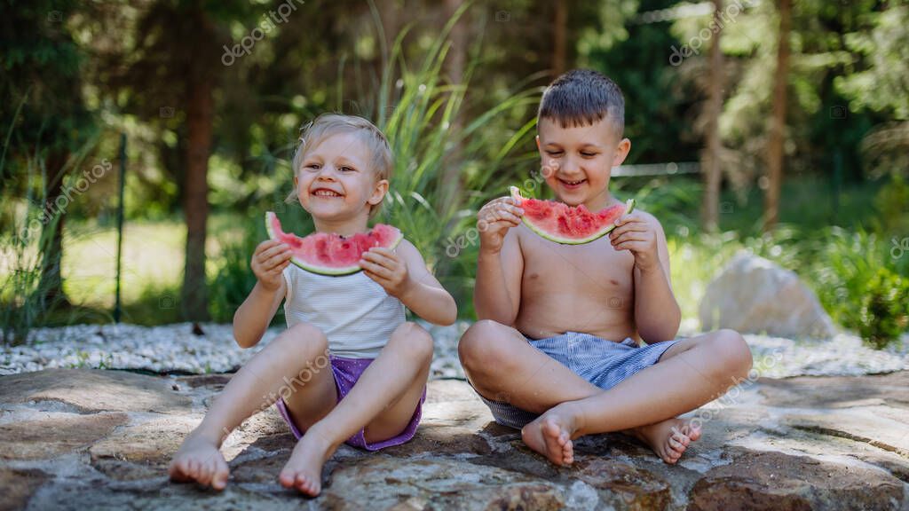 Little chidren sitting near a lake and eating watermelon on hot sunny day during summer vacation.