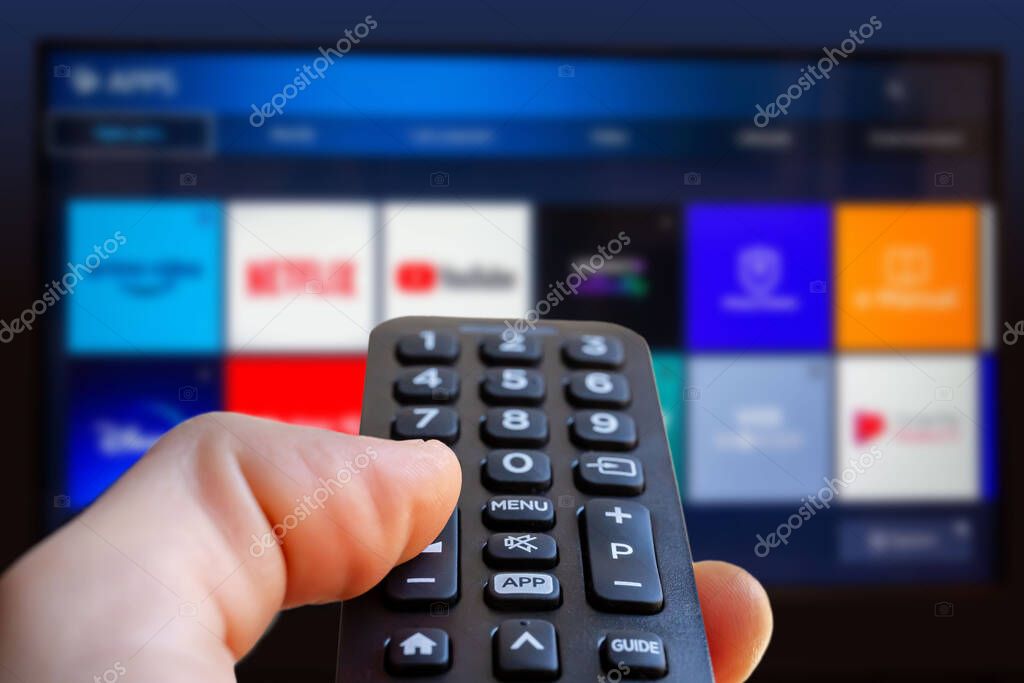 A man is holding the remote control of a smart TV with a television screen in the background with some blurry video streaming service app icon