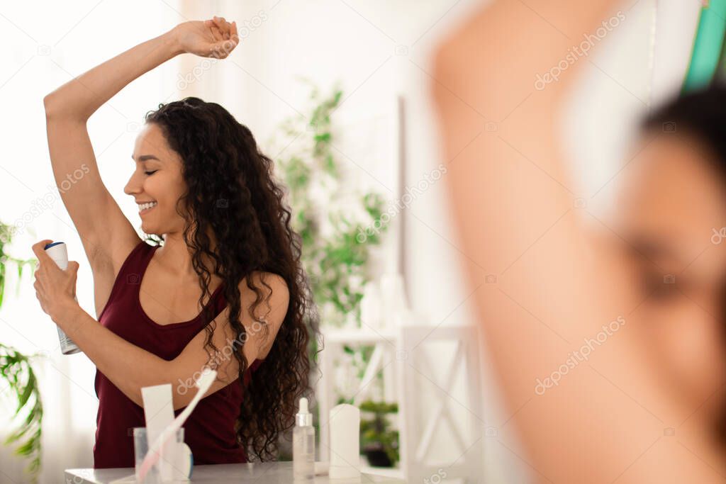 Lovely young woman using spray deodorant on her armpit near mirror at home, free space. Attractive millennial female applying sweat protection in summer. Hygiene and skin care concept