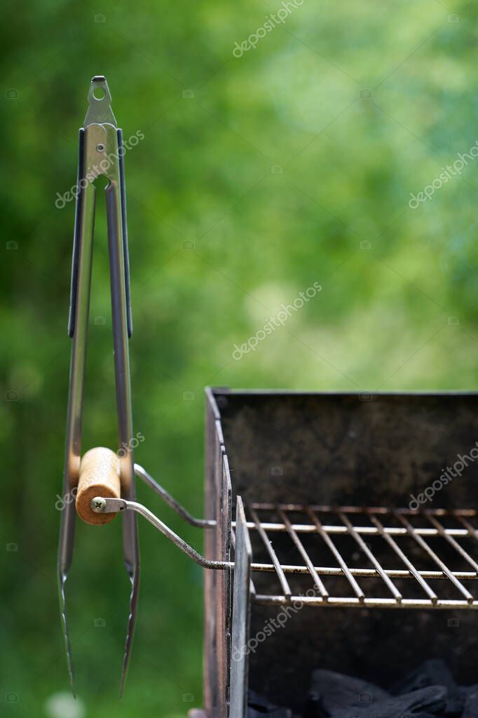 Barbecue tongs (Grillzange) on a wooden handle from a charcoal grill with dirty rust. Green nature in the background. View from the front.