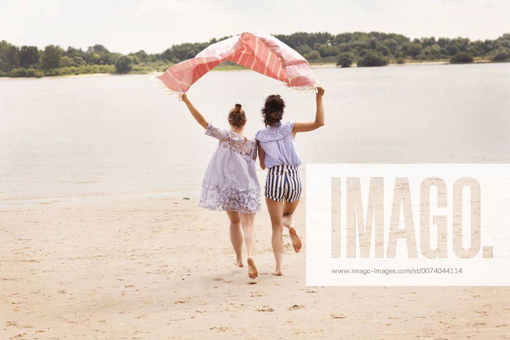 Youniverse - Young Holidays Back view of two friends running side by side on the beach holding cloth over their heads model released Symbolfoto TSFF000115