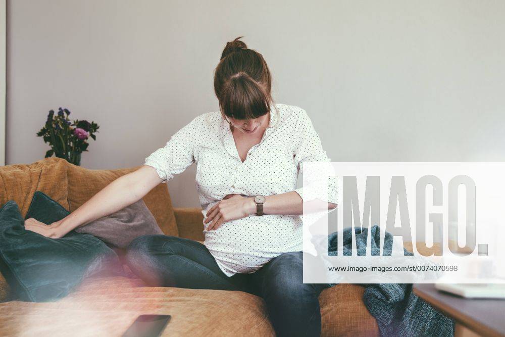 Expectant mother timing her contractions while sitting on couch at home model released Symbolfoto property released MFF03389