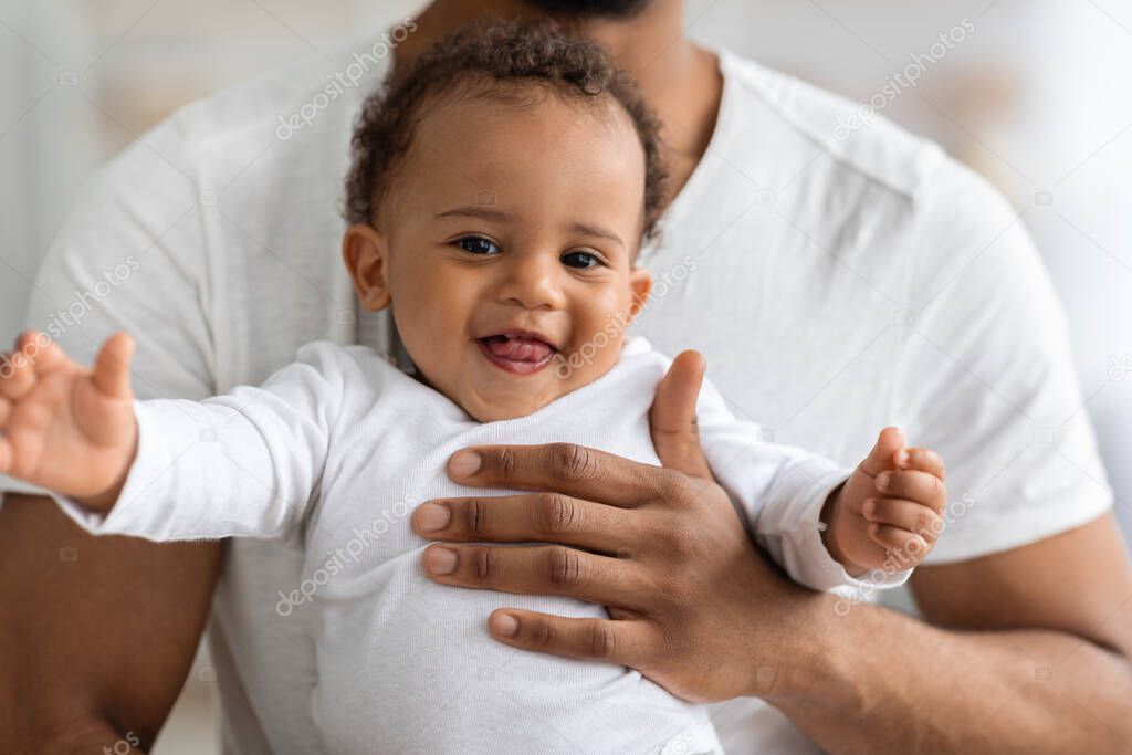 Closeup Portrait Of Adorable Black Baby Boy Relaxing In Fathers Hands, Cute African American Infant Child Smiling And Looking At Camera, Enjoying Spending Time With His Daddy, Cropped Image